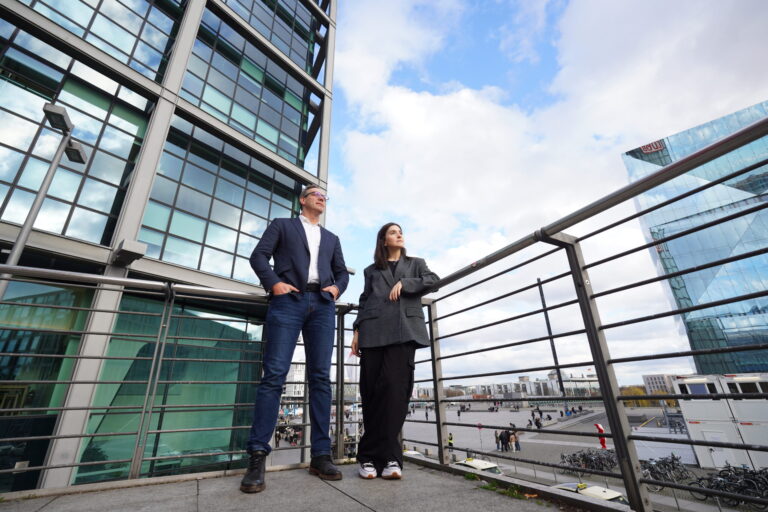 Man and woman standing confidently on a balcony in front of modern glass buildings, overlooking a busy city square